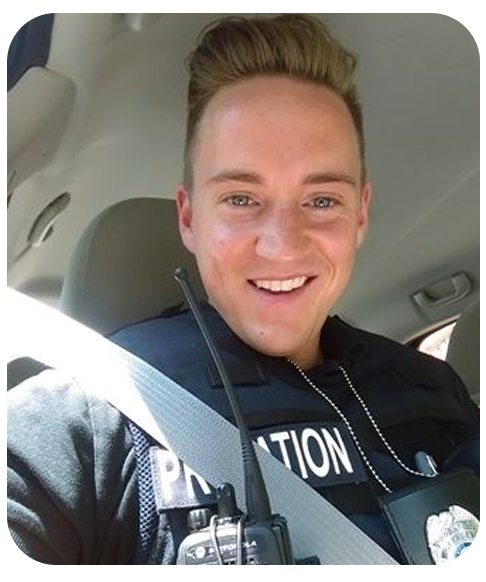 A selfie of Conrad Beauchamp in a vehicle wearing a tactical vest, badge, dogtags, and radio. He is smiling and looking at the camera.