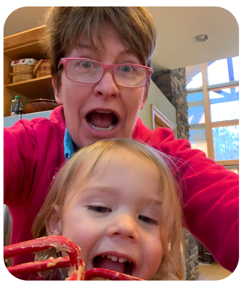 A playful selfie of Jennifer Moser and a toddler-age girl making funny faces in a kitchen. The child licks a whisk.