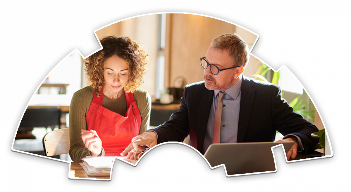 A man in a suit with a laptop points to something on a paper in front of a woman wearing a red apron. They are sitting next to each other.