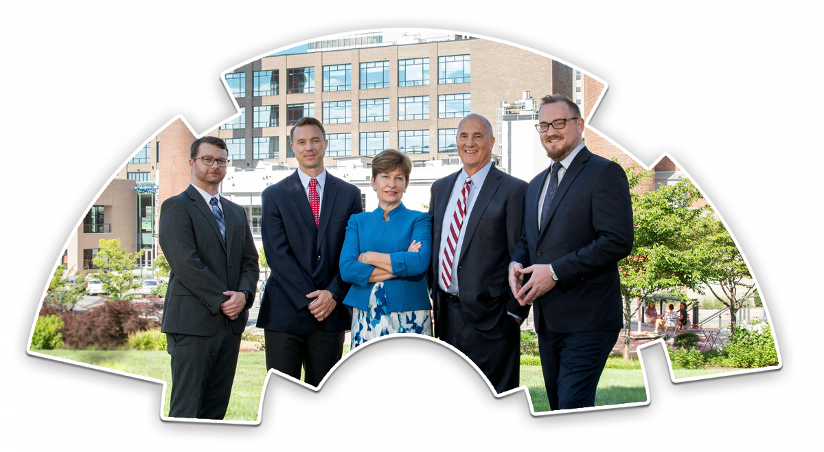 Four men and one woman stand on a grassy patch with part of a city skyline behind them. They are smiling and dressed professionally.