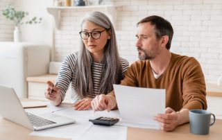 A woman and man look concerned as they sit together at a desk in a home, holding papers and examining a computer screen.