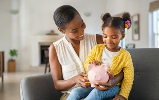 A woman shows a young girl how to save money in a piggy bank while holding her on her lap.