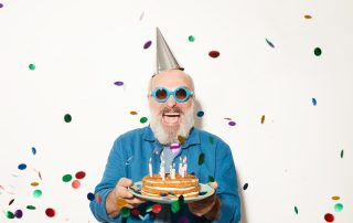 A smiling man celebrates his 59.5 birthday wearing a party hat and sunglasses, holding cake, and surrounded by confetti.