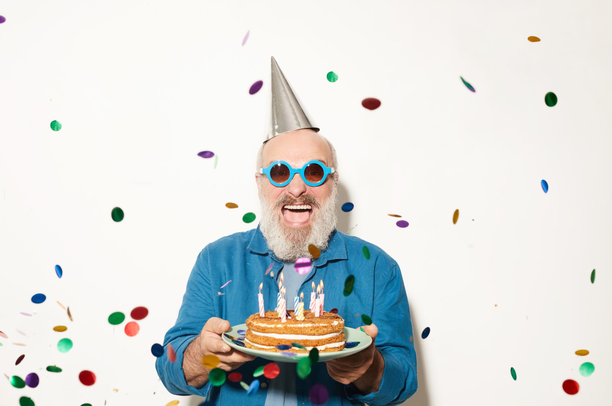 A smiling man celebrates his 59.5 birthday wearing a party hat and sunglasses, holding cake, and surrounded by confetti.