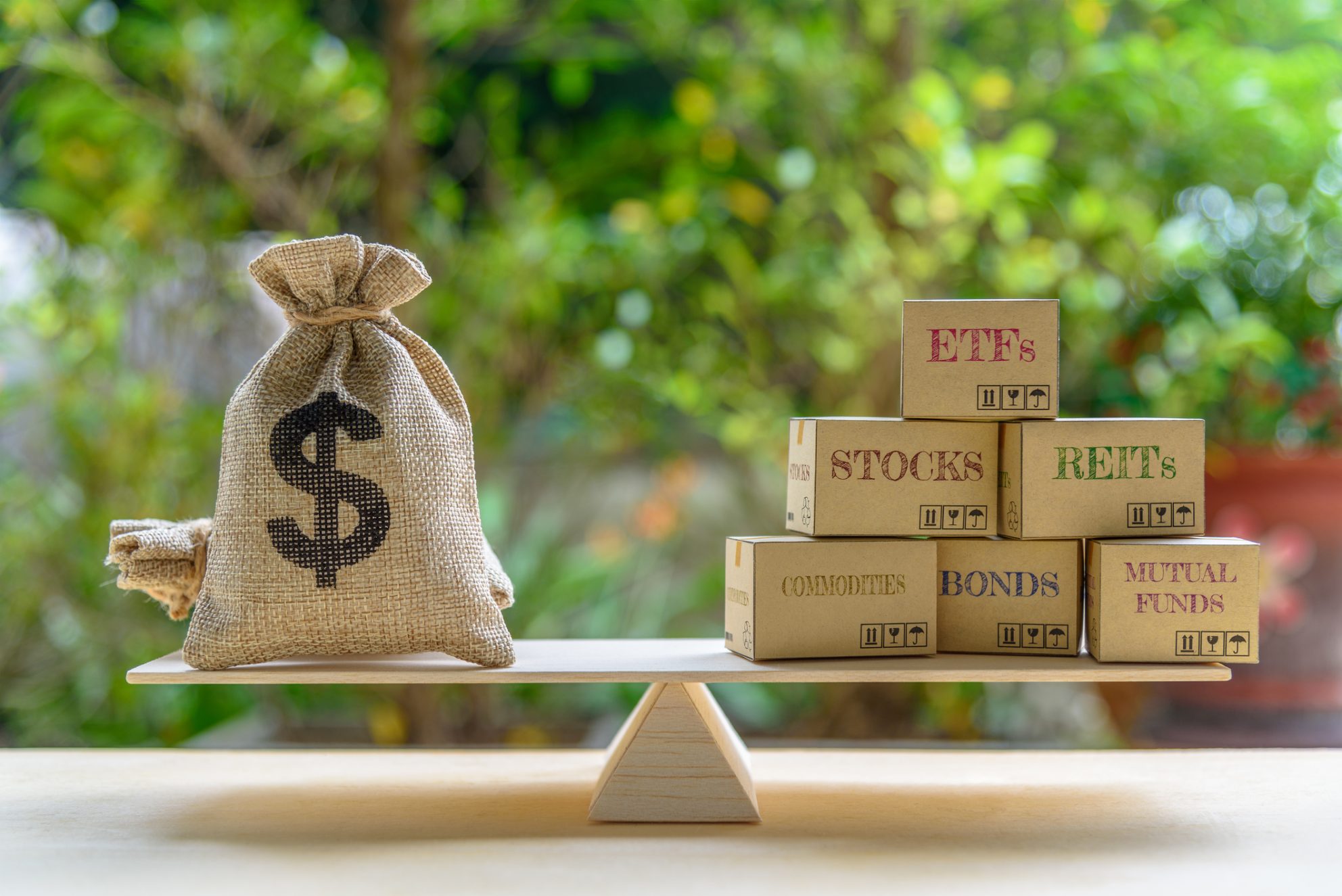 A balanced scale holds a burlap sack with a dollar sign on the left and several boxes on the right, which read ETFs, Stocks, REITs, Commodities, Bonds, and Mutual Funds.