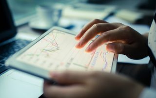 A person's hands hold a bright tablet with colorful graphs on them. The graph lines overlap and intersect. In the background there's a desk with a computer, coffee cup, and papers.