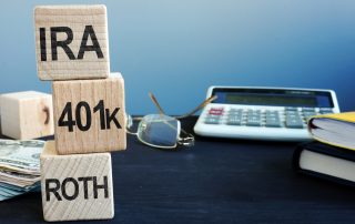 Several items sit on a desk, including a large calculator, glasses, books, and papers. In the foreground, there are three blocks stacked vertically. From top to bottom, they read: IRA, 401K, and ROTH.