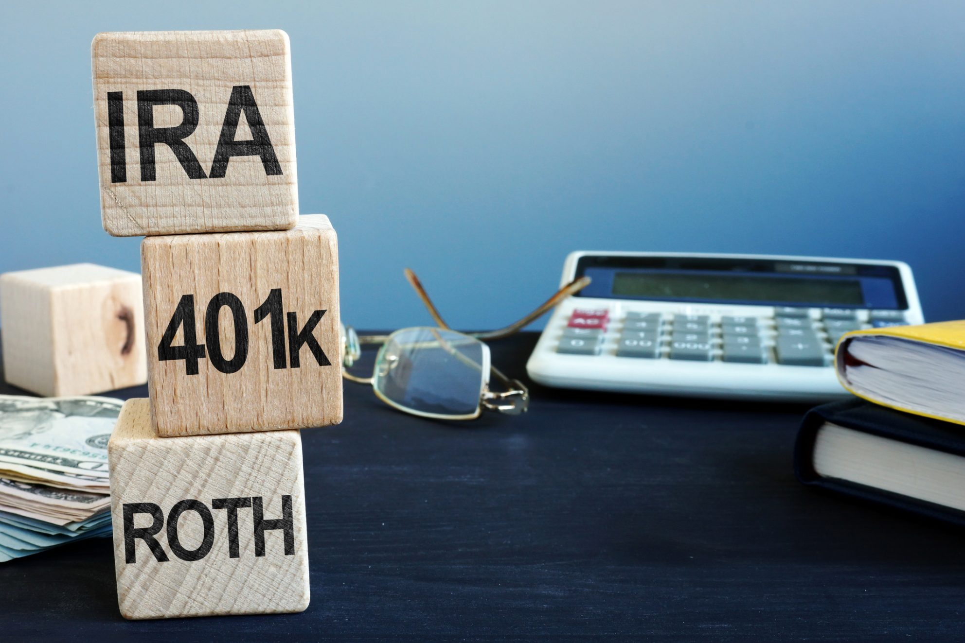 Several items sit on a desk, including a large calculator, glasses, books, and papers. In the foreground, there are three blocks stacked vertically. From top to bottom, they read: IRA, 401K, and ROTH.