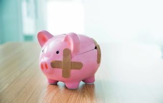 A pink piggy bank stands on a table in a bright room. It is cracked and held together using three brown band-aids.