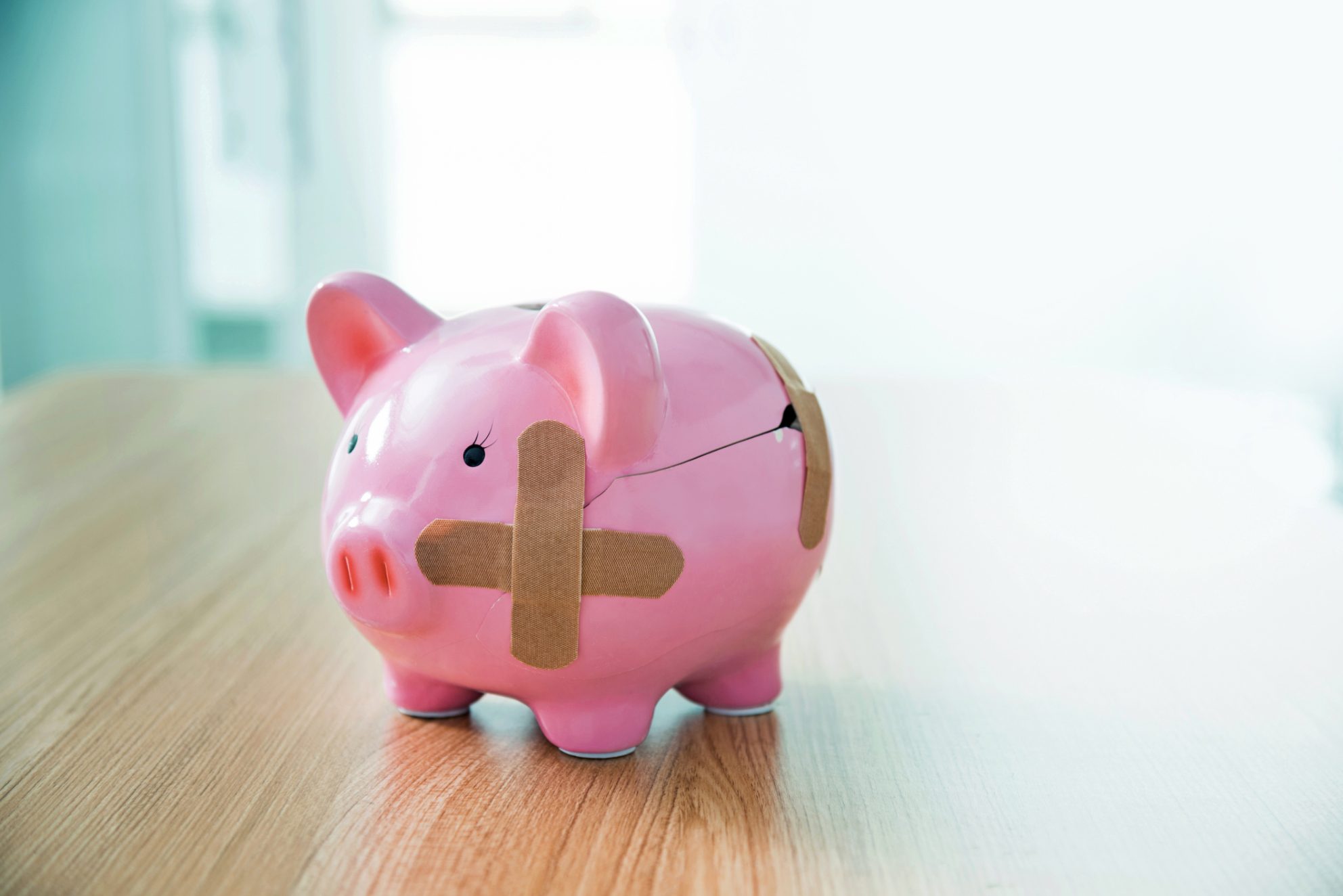 A pink piggy bank stands on a table in a bright room. It is cracked and held together using three brown band-aids.