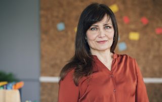 A professional adult woman smiles slightly and stands in front of a bulletin board with small colorful papers on it.